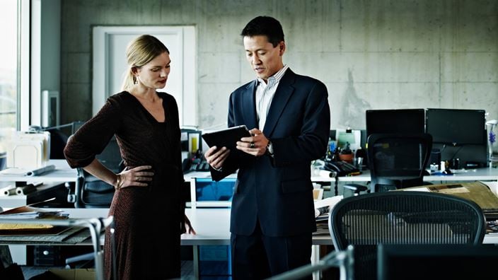 While viewing a digital tablet, a businesswoman and businessman stand in a modern office discussing how to leverage Crowe AI governance framework to securely utilize their proprietary AI platform for tasks like document review