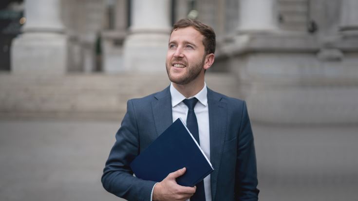 Grant manager wearing a navy blue suit, carrying a blue folder looks off into the distance, thinking how to maximize his funds