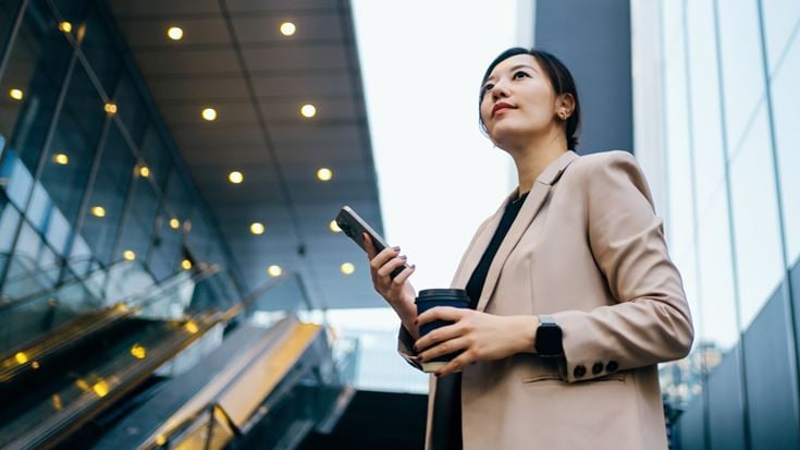 Professional holding a phone and coffee outside a modern office, representing banking transformation through technology.