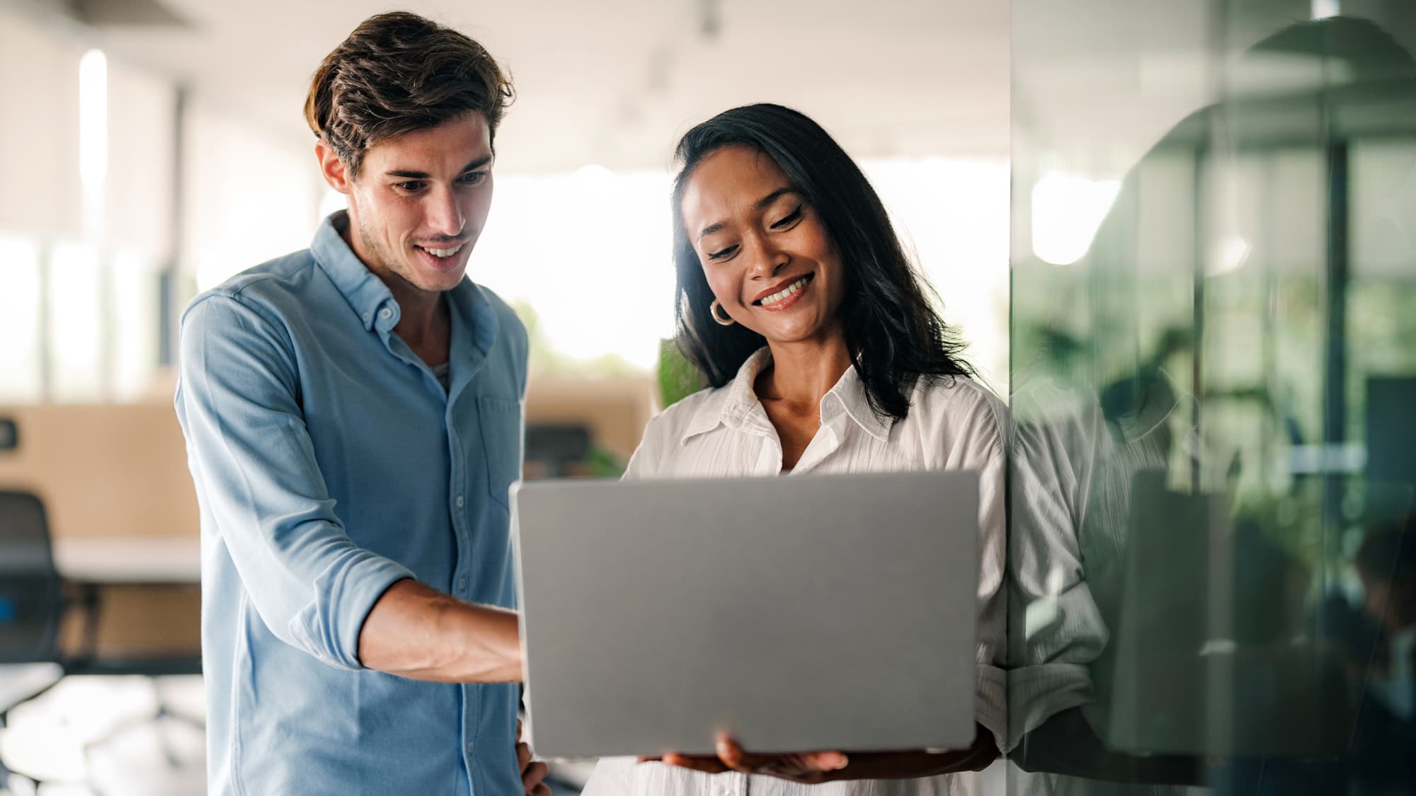 Two professionals smiling while reviewing information together on a laptop in an office setting.