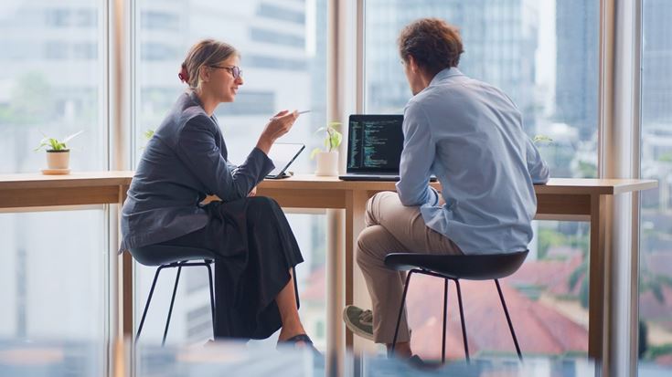 Two professionals discuss AI governance strategy at a desk with a laptop displaying code.
