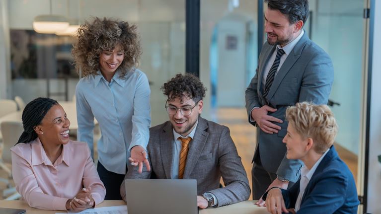 Diverse business professionals collaborating around a laptop during a team meeting in a modern office, discussing ideas and project strategy.