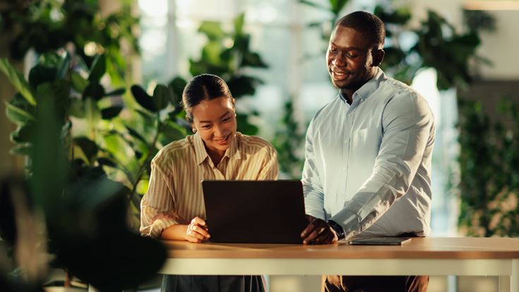 Two banking professionals collaborating at a laptop in a bright, plant-filled office.