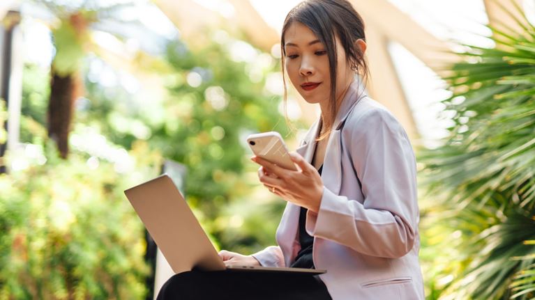 Professional reviews information on phone and laptop outdoors, representing efficient, tech-enabled risk management in financial services.