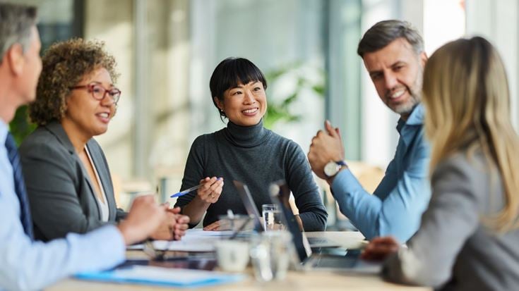 A diverse group of five professionals sitting around a conference room table, engaged in an active business meeting.