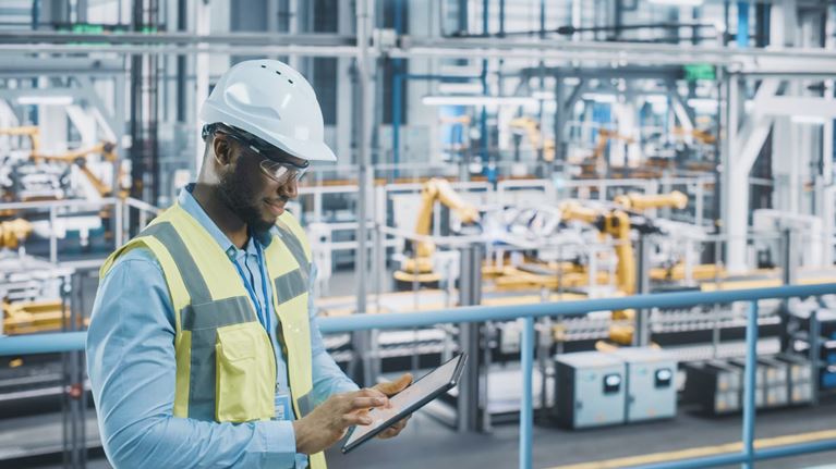 Manufacturing professional using a tablet on a factory floor with automated equipment and industrial robotics.