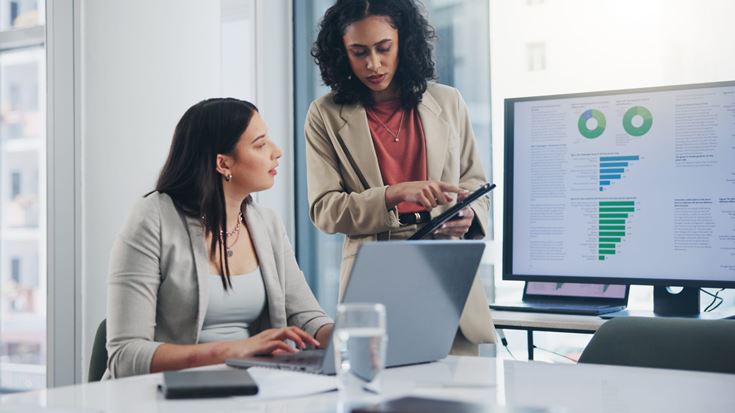 Two business professionals collaborate in a modern office, reviewing analytics dashboards with charts and graphs displayed on a large monitor while one points to insights on a tablet and the other works on a laptop—illustrating how teams interpret data to guide AI and analytics strategy.
