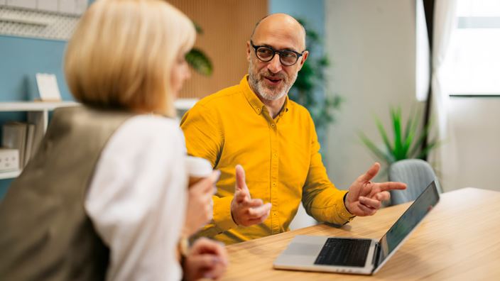 Two professionals discuss work across a conference table with a laptop during an office meeting.
