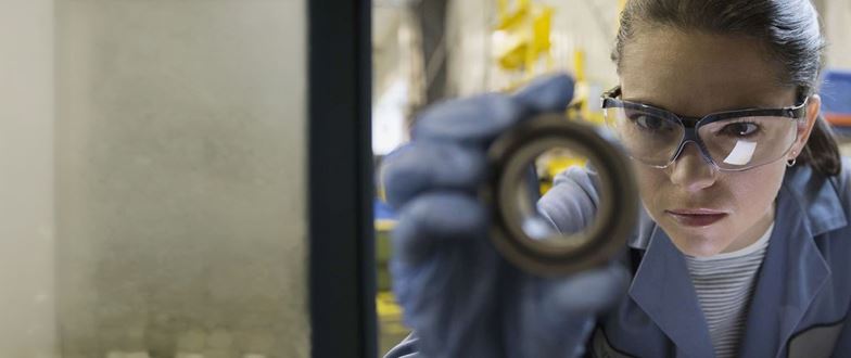 Metals industry worker inspects a metal component while wearing protective eyewear and gloves.