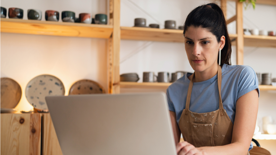 Woman in apron working on a laptop, reading Payday superannuation updates in a pottery studio, with shelves of pottery and ceramics in the background.