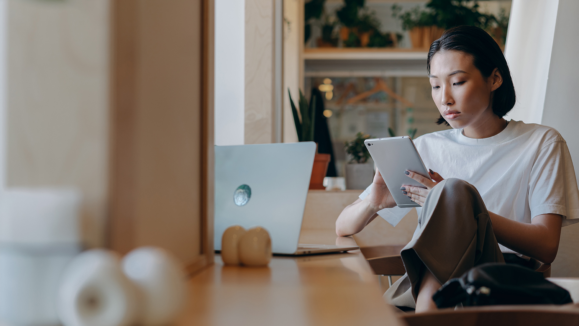 Woman looking at her tablet while her laptop is on the table in front of her.