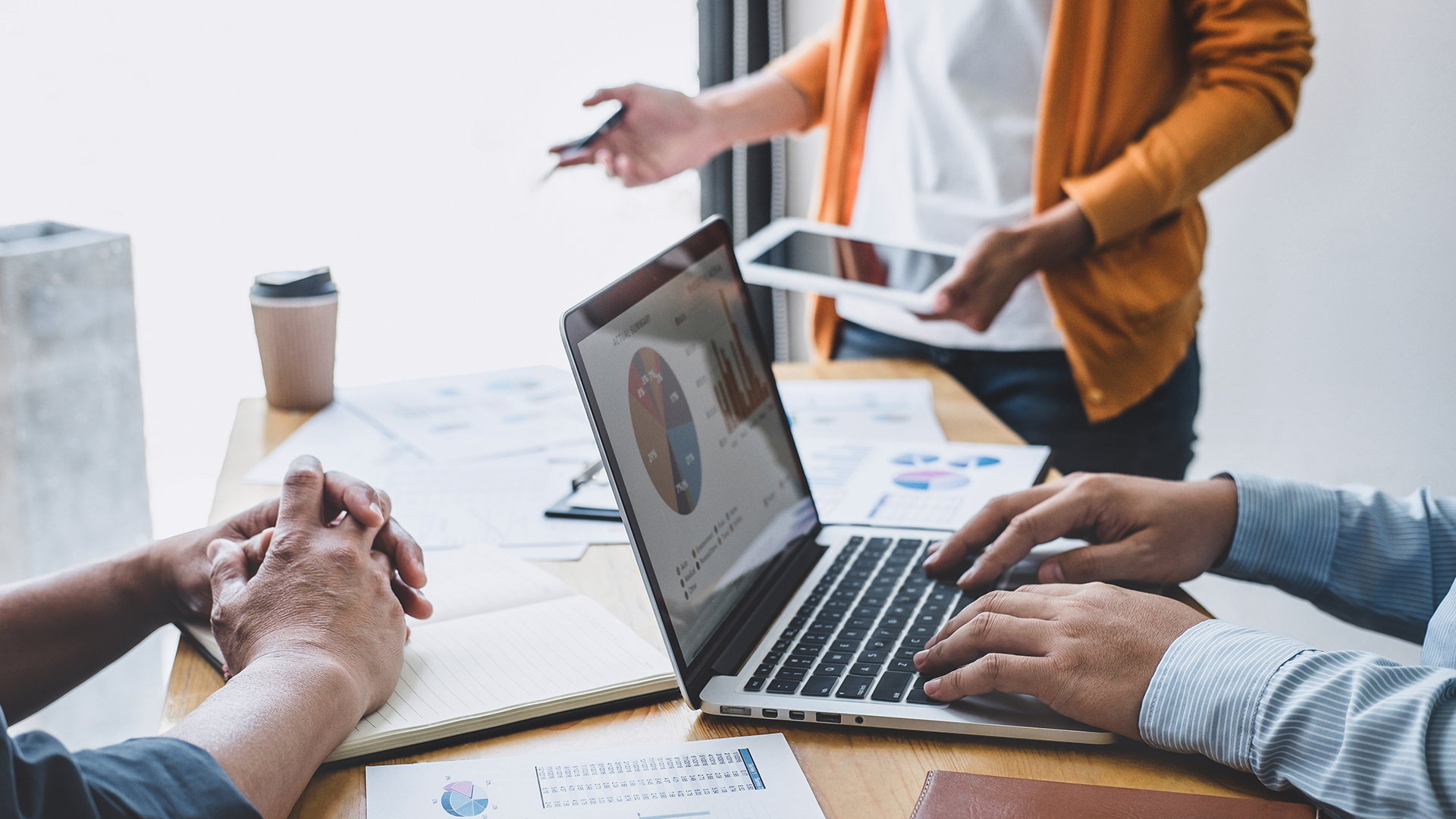 Close up of three people.  One has a laptop open with a pie graph displayed, one is holding a tablet.  The table is covered with spreadsheets and graphs.