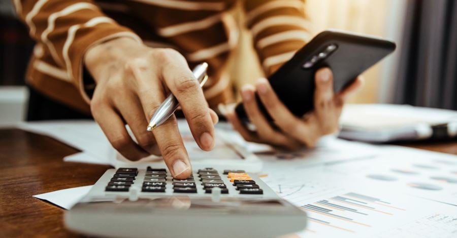 A person holding a pen and punching figures into a calculator in the foreground, while holding their phone