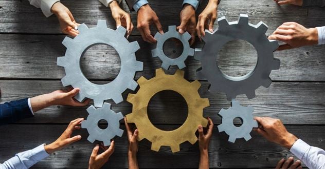 A cog mechanism is being held together by hands of multiple people reaching in from outside the frame