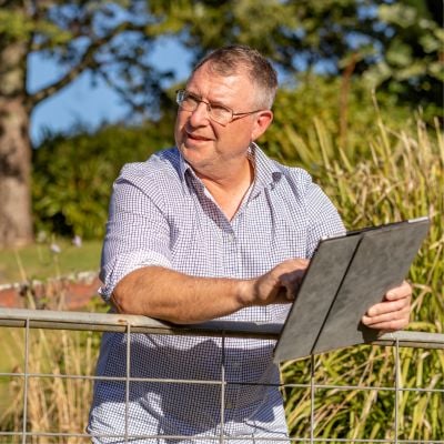Man standing up leaning on a fence, holding a tablet with trees and long grass surrounding him.